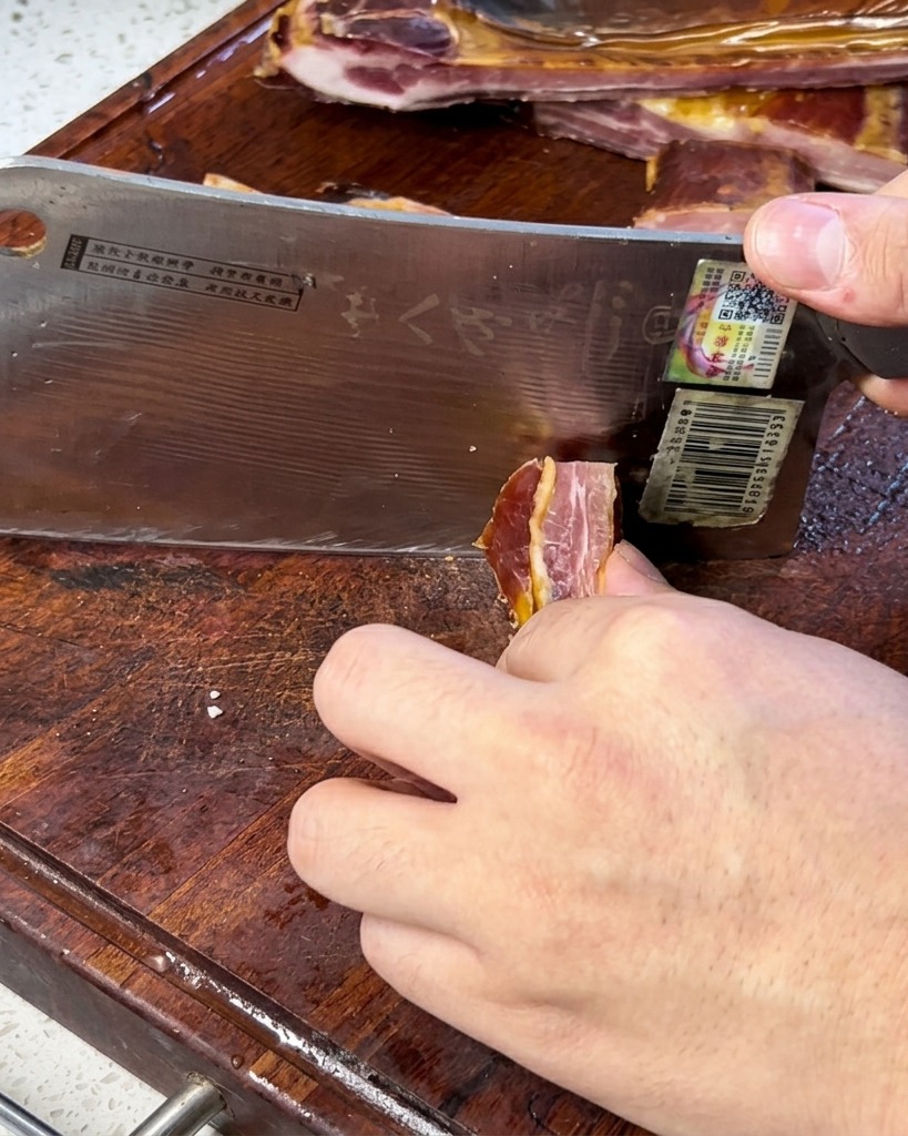 A close-up shot of a chef cutting thin slices of cured pork belly or rib meat using a broad kitchen cleaver on a wooden cutting board.