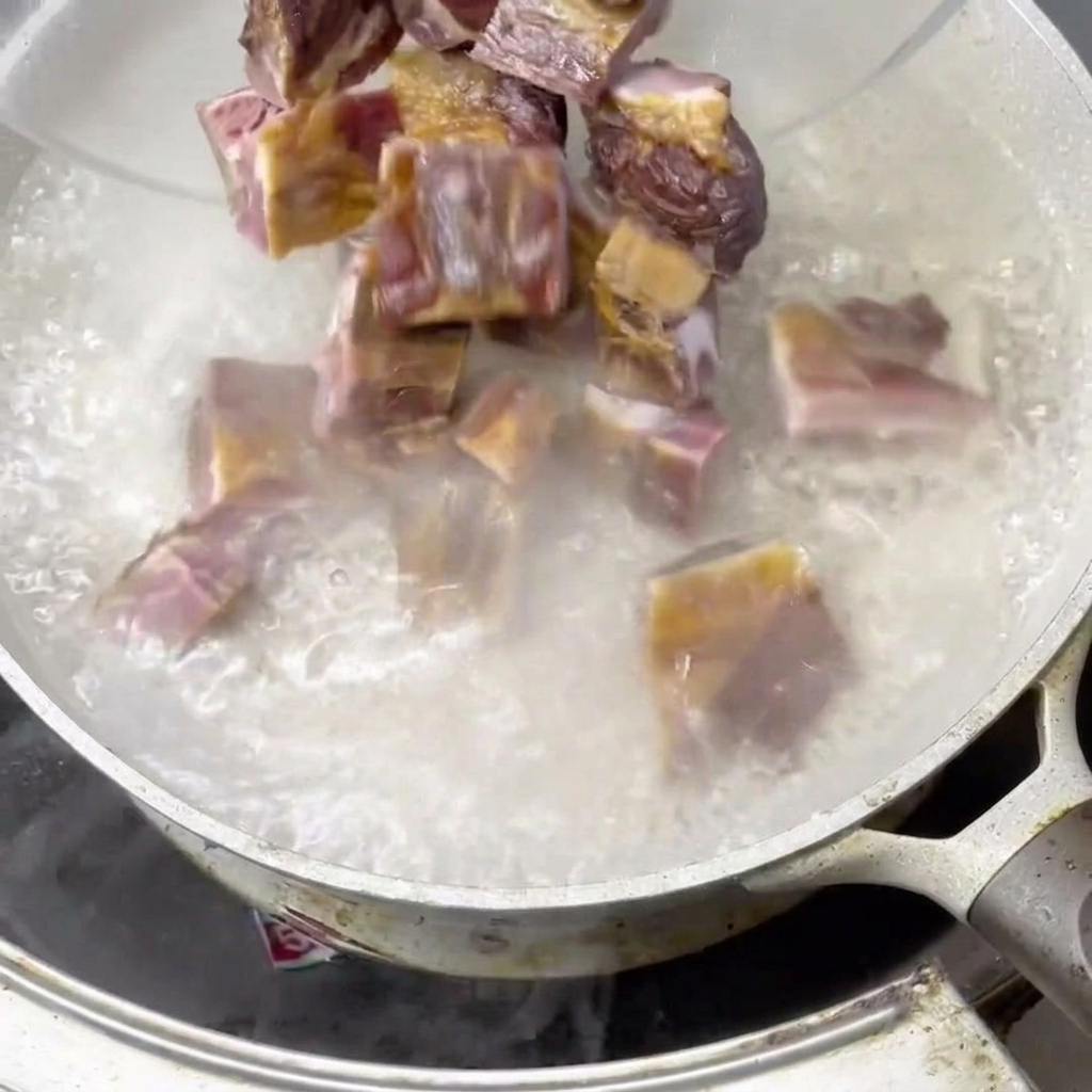 Pieces of cured pork ribs being dropped from a bowl into a pot of rapidly boiling water, creating steam.