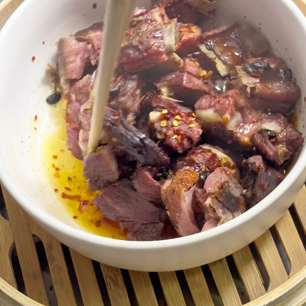 A pair of chopsticks mixing steamed pork ribs with red chili flakes and black beans in a white bowl inside a bamboo steamer basket.