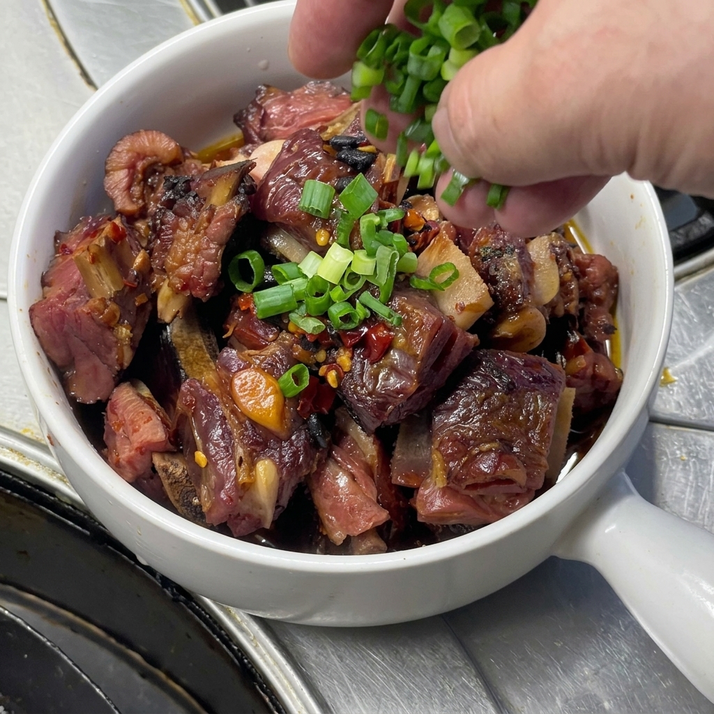 Freshly chopped green onions being sprinkled from a hand onto a white handled bowl filled with glossy steamed pork ribs, garlic cloves, and chili.