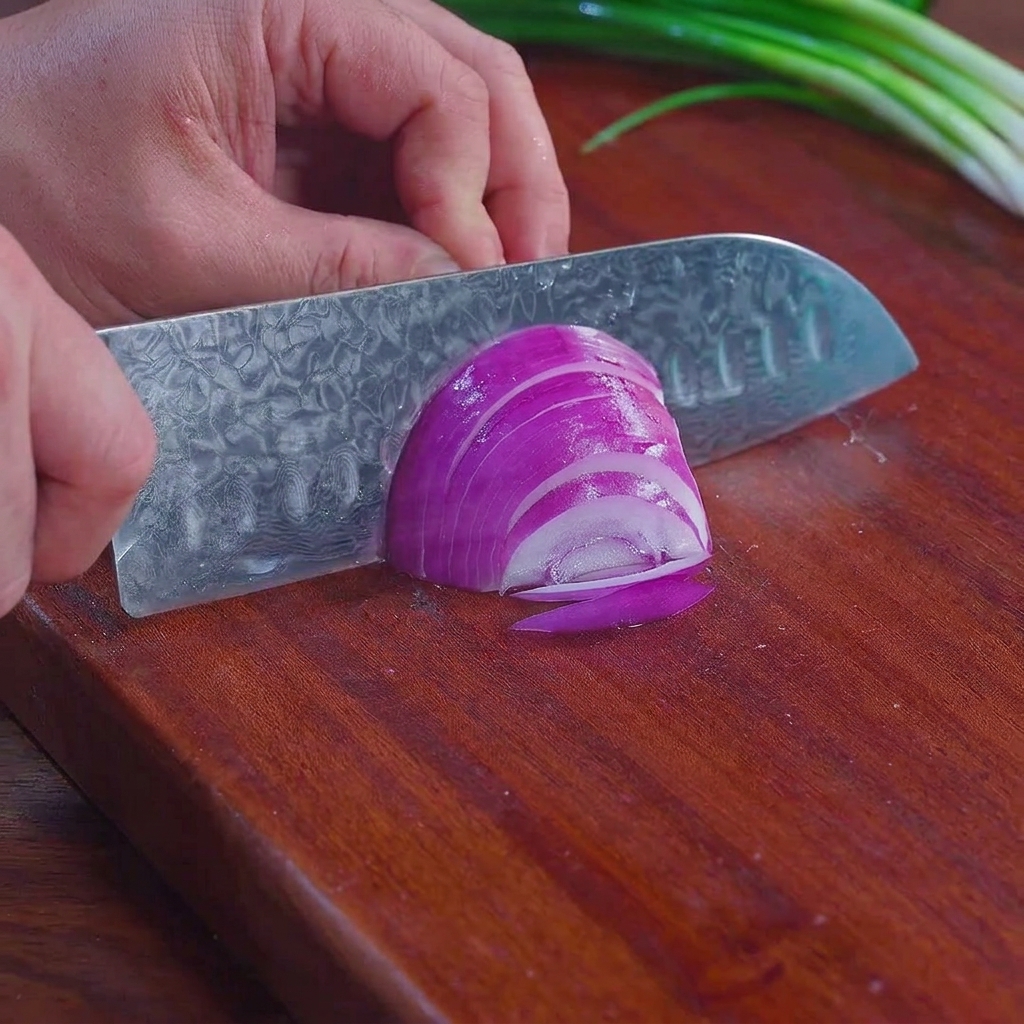 A chefs knife slicing a purple red onion into thin shreds on a wooden cutting board with green onions visible in the background.