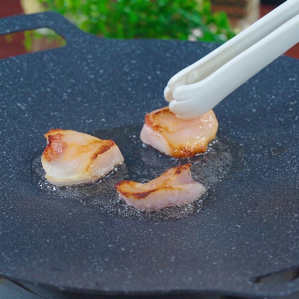 White paw-shaped tongs rubbing pieces of pork fat across a hot, seasoned black grill pan to render lard.