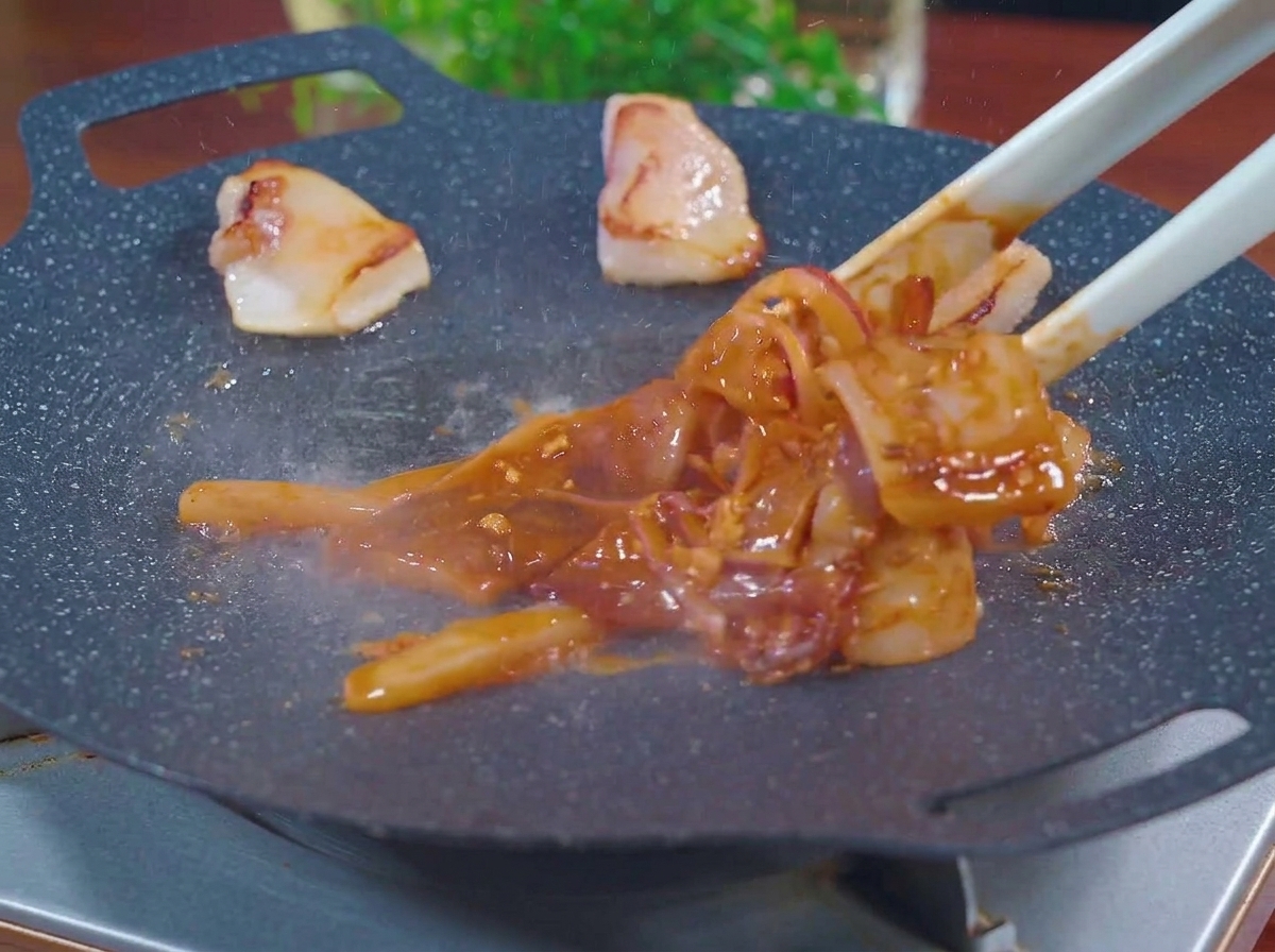 Tongs placing marinated red squid pieces and onion slices onto a hot, smoking black grill pan alongside rendered pork fat chunks.