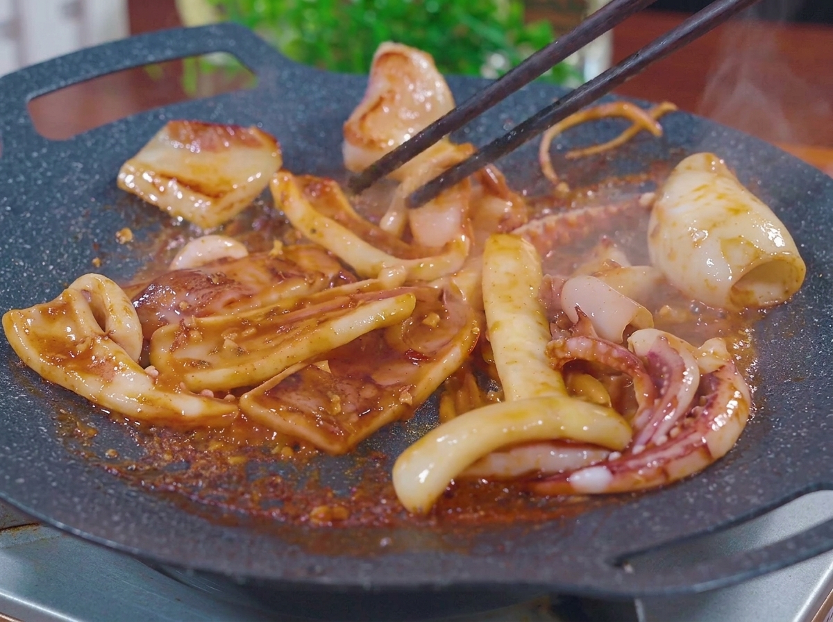 Squid pieces being stir-fried with chopsticks on a flat, hot griddle pan, sizzling in a thick, bubbly reddish-brown barbecue marinade with steam rising.