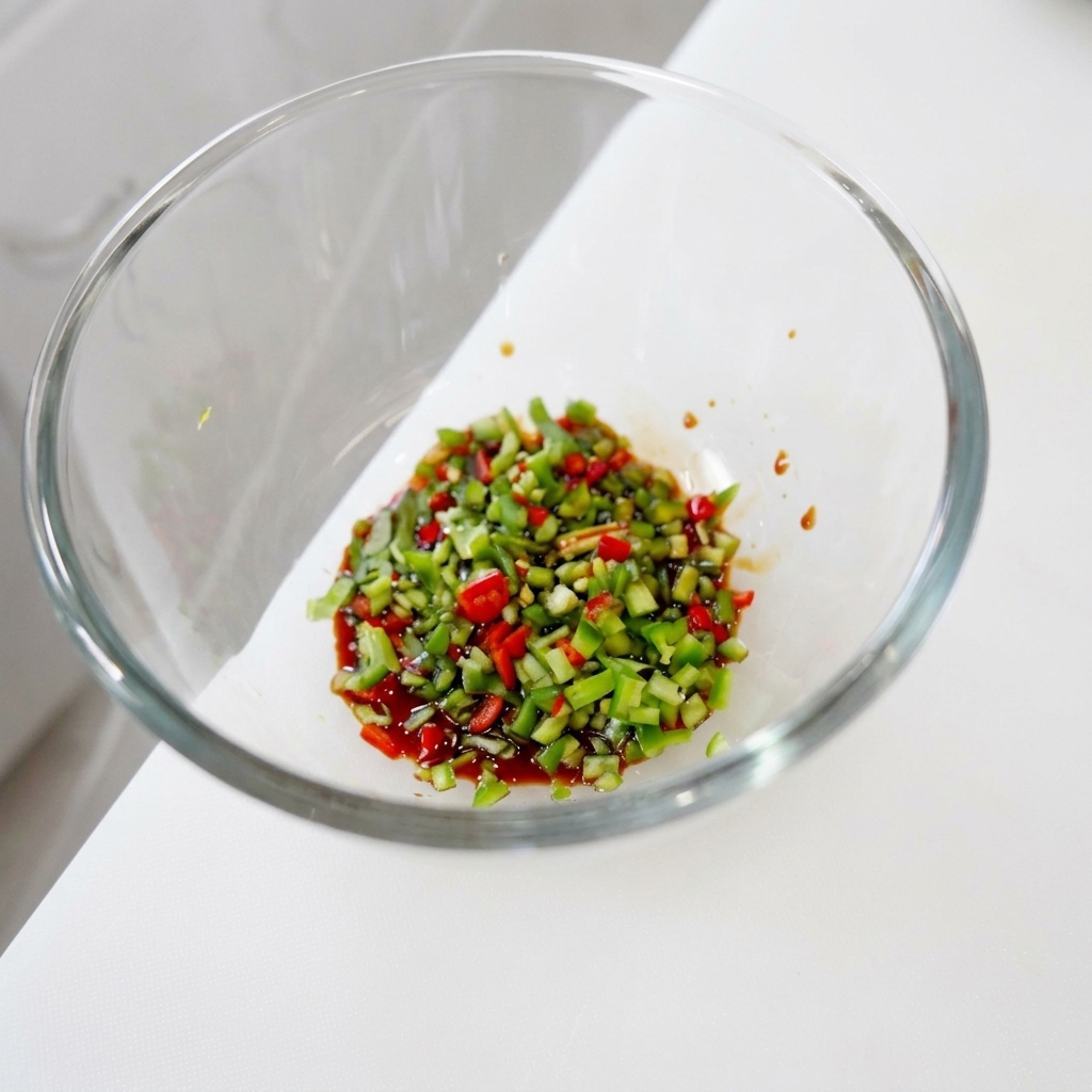 A vibrant mixture of finely minced green and red chilies and cilantro soaking in a dark, glossy liquid seasoning base within a glass bowl.