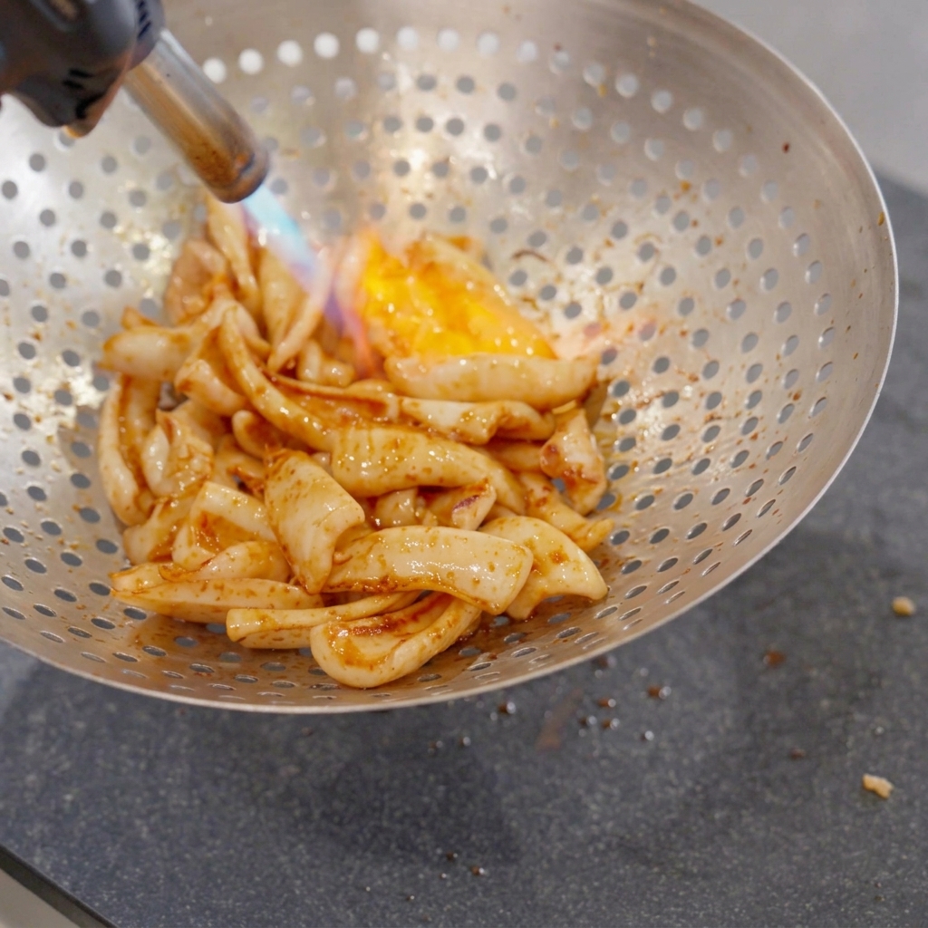 Fried squid pieces inside a large metal perforated strainer being blasted by a blue and orange flame from a hand-held kitchen blowtorch.