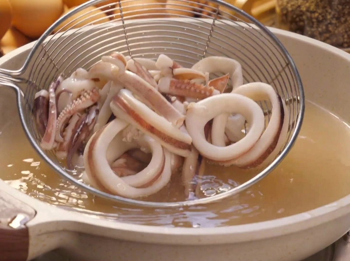 A wire mesh strainer lifting blanched squid rings out of a pot of boiling water.