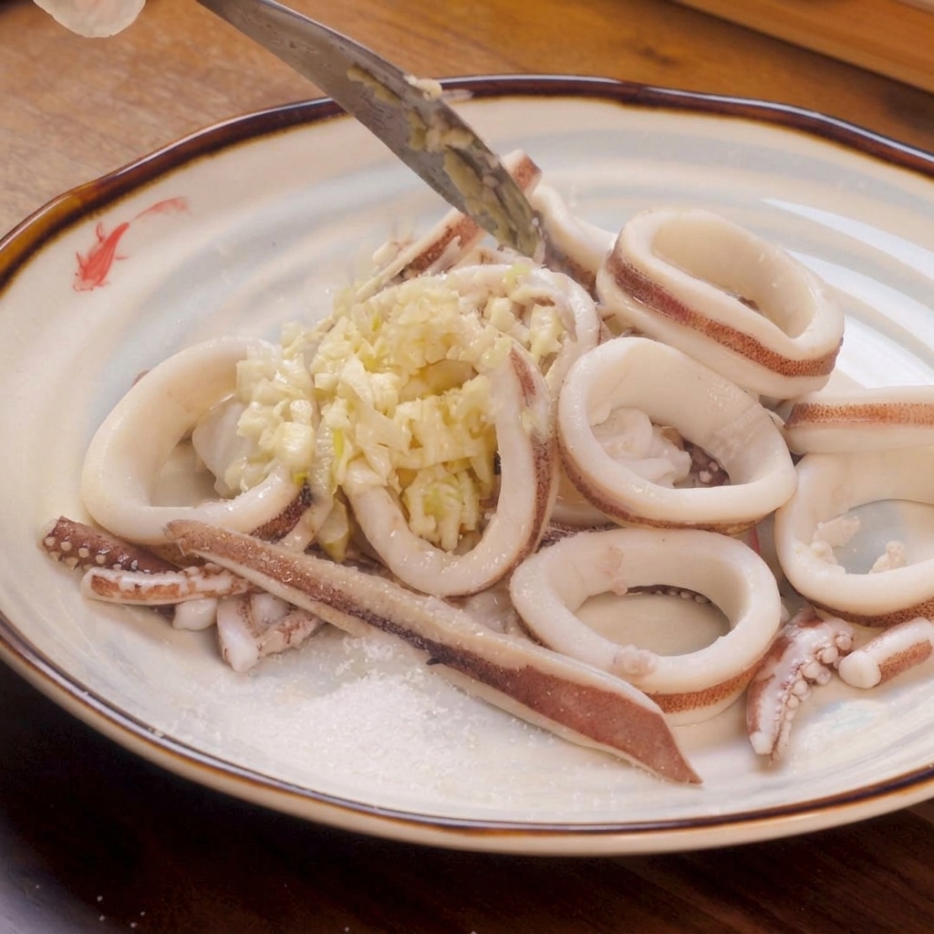 Blanched white squid rings arranged on a plate, topped with a heap of minced garlic and a sprinkle of salt and sugar on the side.