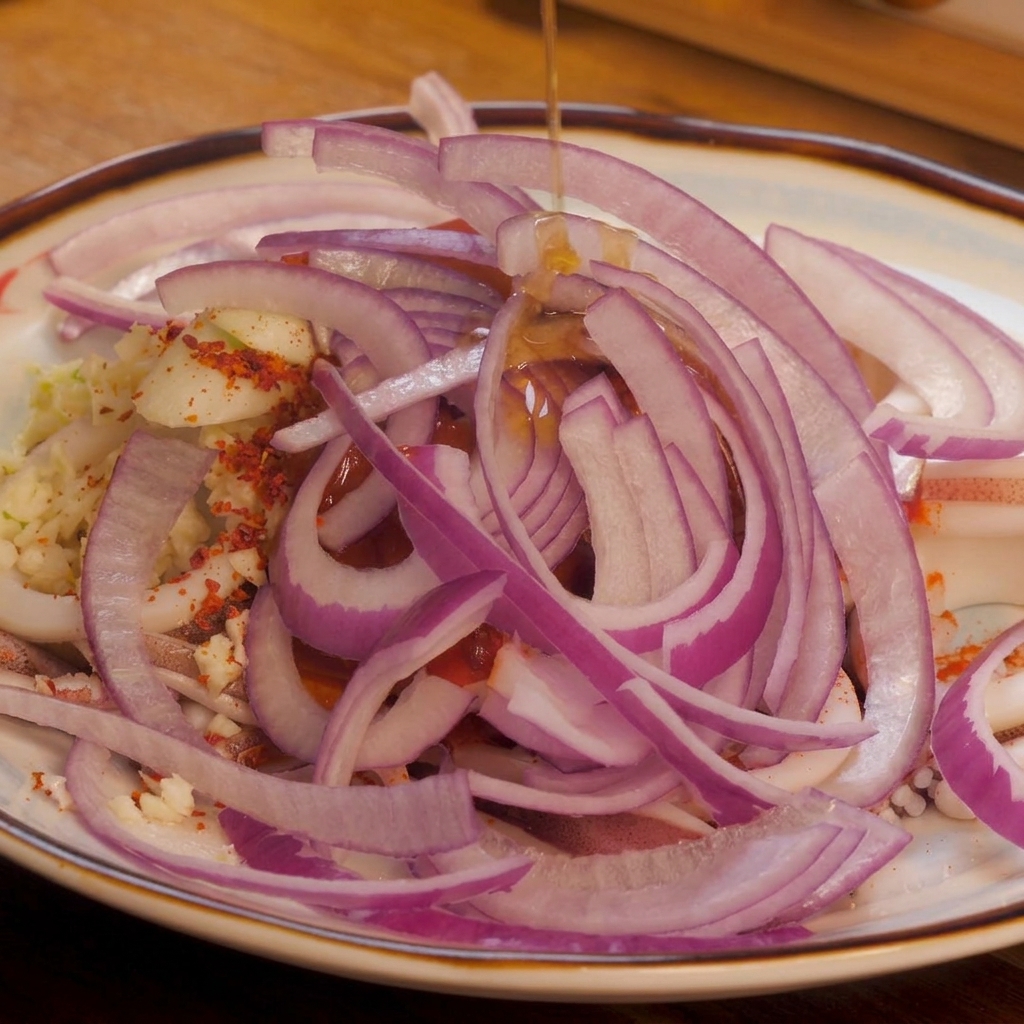 A stream of clear sesame oil being drizzled from above onto a colorful pile of sliced red onions, chili powder, and squid.