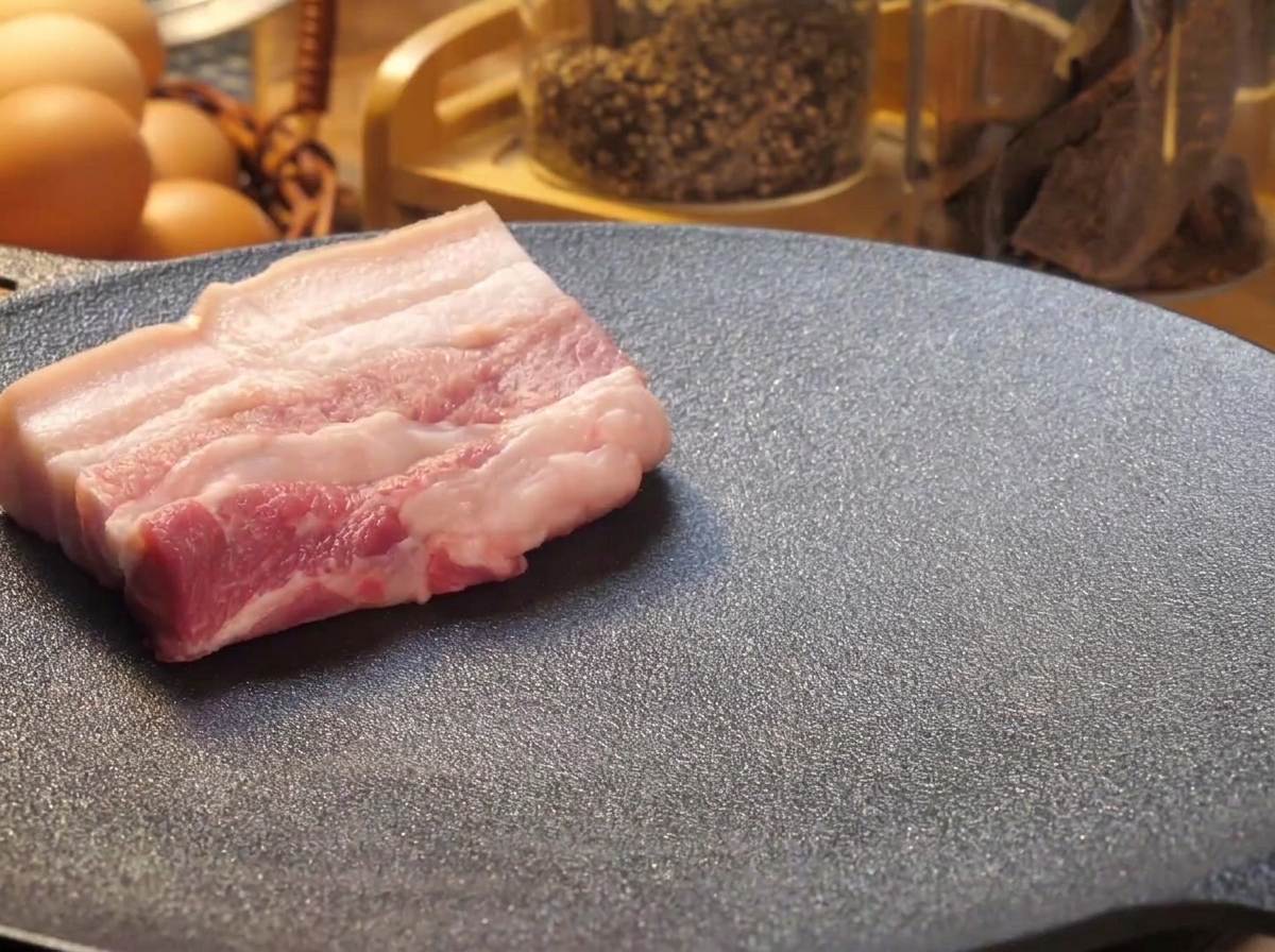 Thick slices of raw pork belly placed onto a hot black griddle pan to sear.