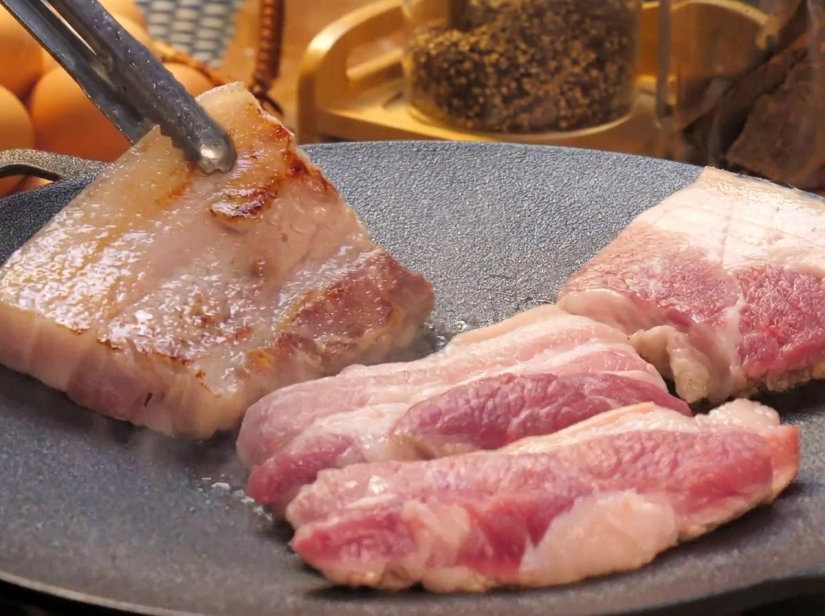 Thick slices of pork belly cooking on a flat griddle pan, with one piece being flipped over by metal tongs to reveal its seared golden surface.