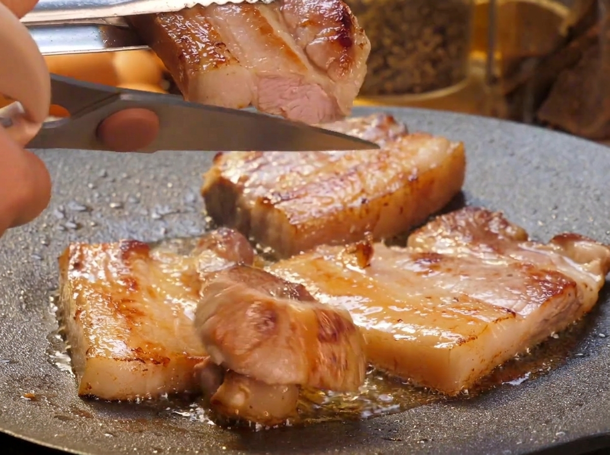 A hand using kitchen scissors to cut thick, seared pork belly slices into smaller strips over a sizzling griddle pan.
