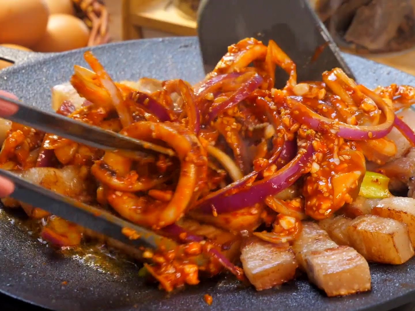 A spatula and tongs stir-frying marinated squid rings and pork belly strips with red onions on a sizzling griddle.