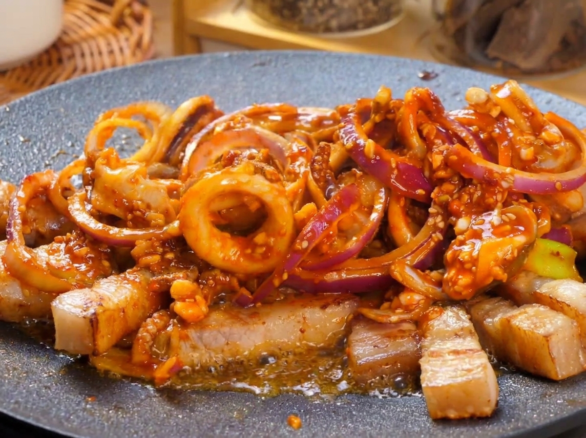 A hot griddle filled with a spicy, red-sauced pork belly and squid stir-fry bubbling vigorously, topped with fresh green pepper garnishes.