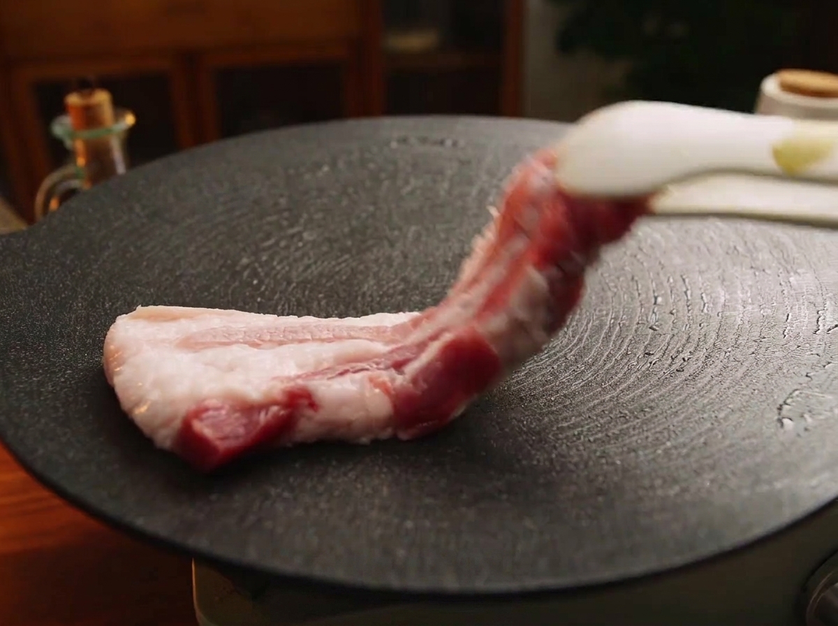 A thick strip of raw pork belly being lowered onto a hot, black iron griddle plate with tongs.