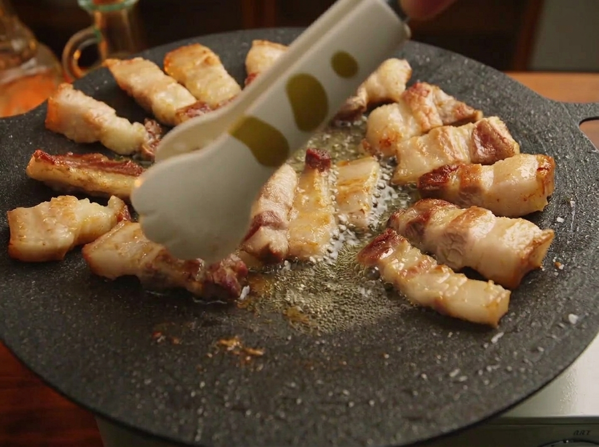 A pair of tongs arranging bite-sized pieces of grilled pork belly in a circular pattern around the rim of a black cast-iron griddle.