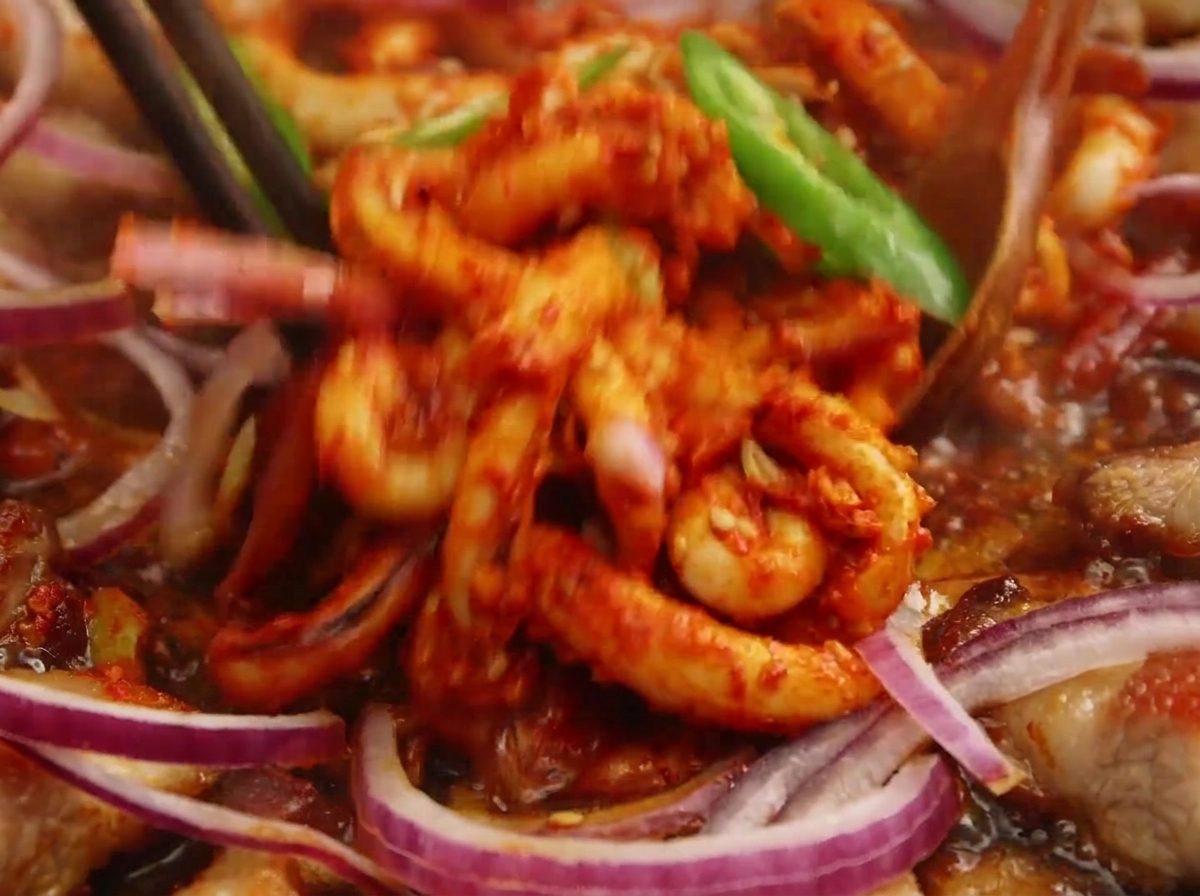 Marinated spicy squid being tossed with chopsticks and a wooden spoon in a pan, surrounded by freshly added red onion rings and green chili slices.