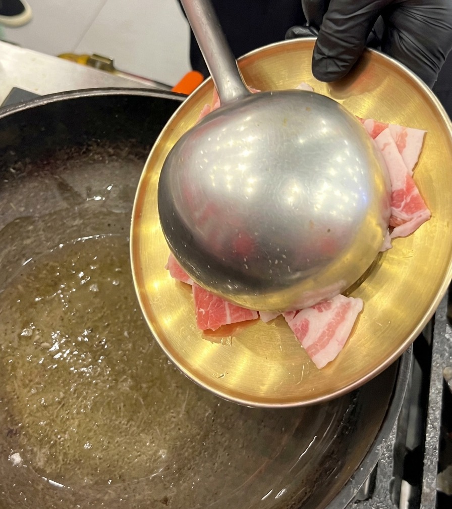 Slices of raw pork belly being transferred from a golden plate into an oiled wok using a large metal ladle.