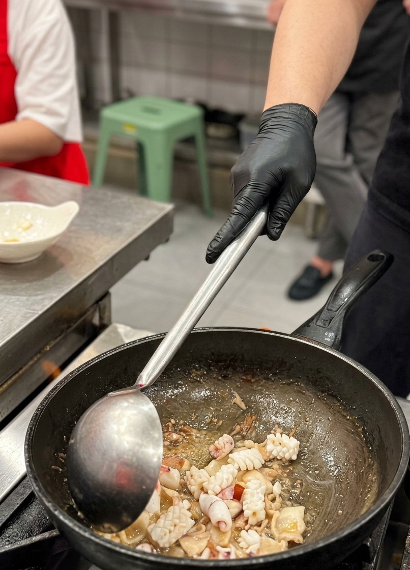 A close-up shot of criss-cross scored squid pieces being stirred into a wok alongside seared pork belly slices using a large metal ladle.