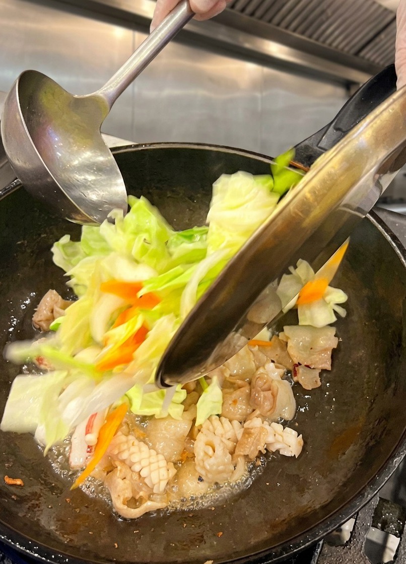 Freshly chopped green cabbage and sliced carrots being poured from a metal plate into a hot wok containing cooked pork belly and squid.