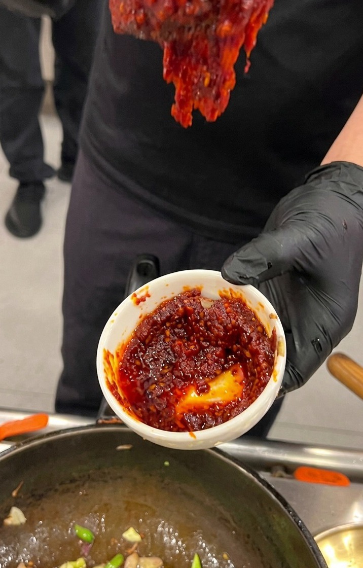 A hand wearing a black glove holding a small white bowl filled with thick, rich red Korean chili paste sauce over a cooking wok.