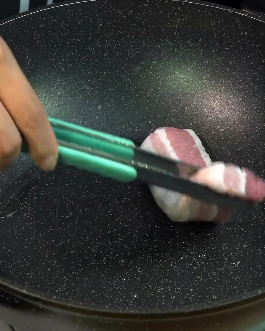 Thick slices of raw pork belly being placed into a black pan using light green tongs to sear the meat.