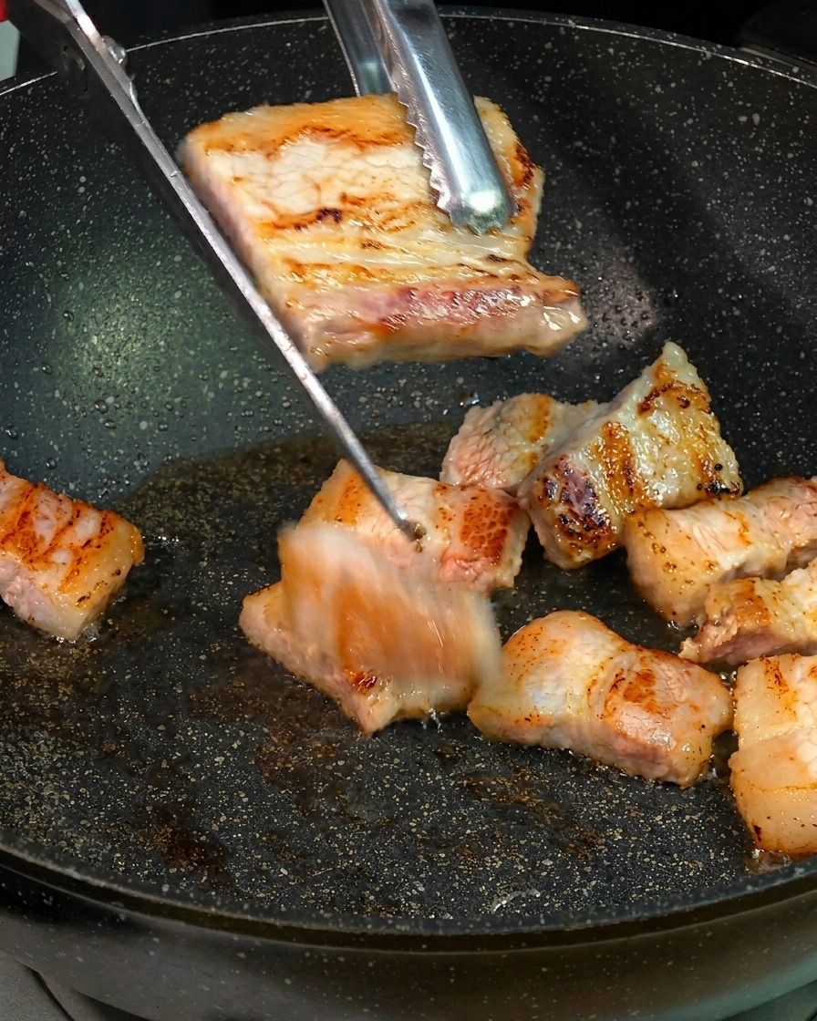 Golden brown seared pork belly slices being cut into smaller bite-sized pieces with kitchen scissors over a frying pan.