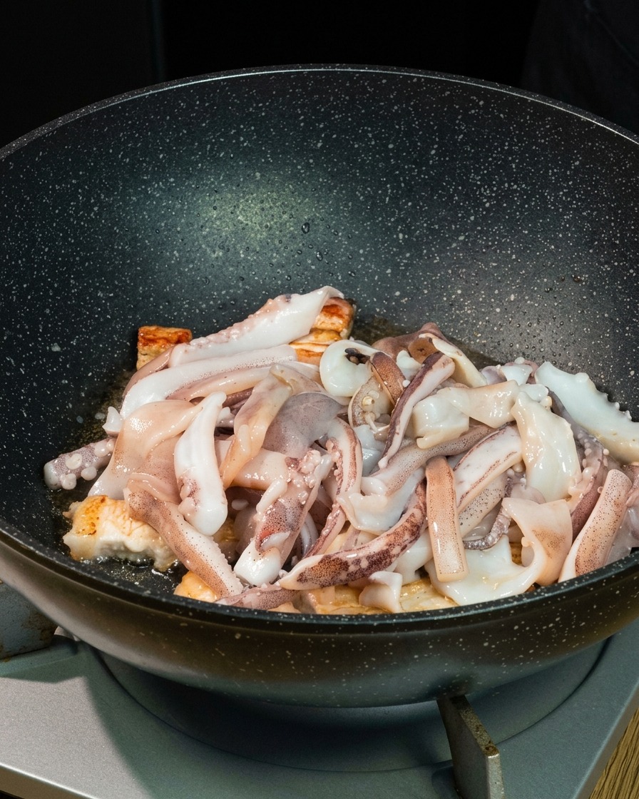 Fresh squid pieces layered over seared pork belly bites inside a deep black wok.