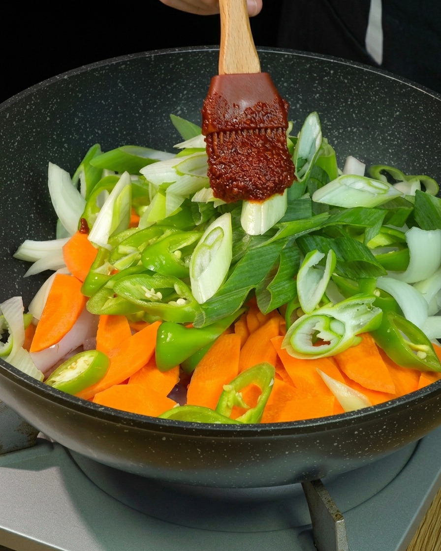 A thick dollop of dark red chili sauce on a wooden spatula being placed over a vibrant pile of sliced carrots, green peppers, onions, and scallions in a pan.