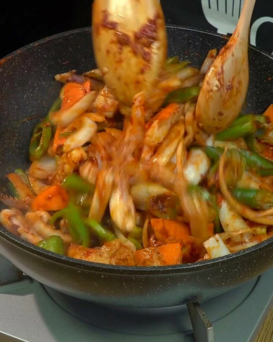 Motion blur of pork belly, squid, and mixed vegetables being vigorously tossed and stir-fried with two wooden spoons in a wok, fully coated in red spicy sauce.