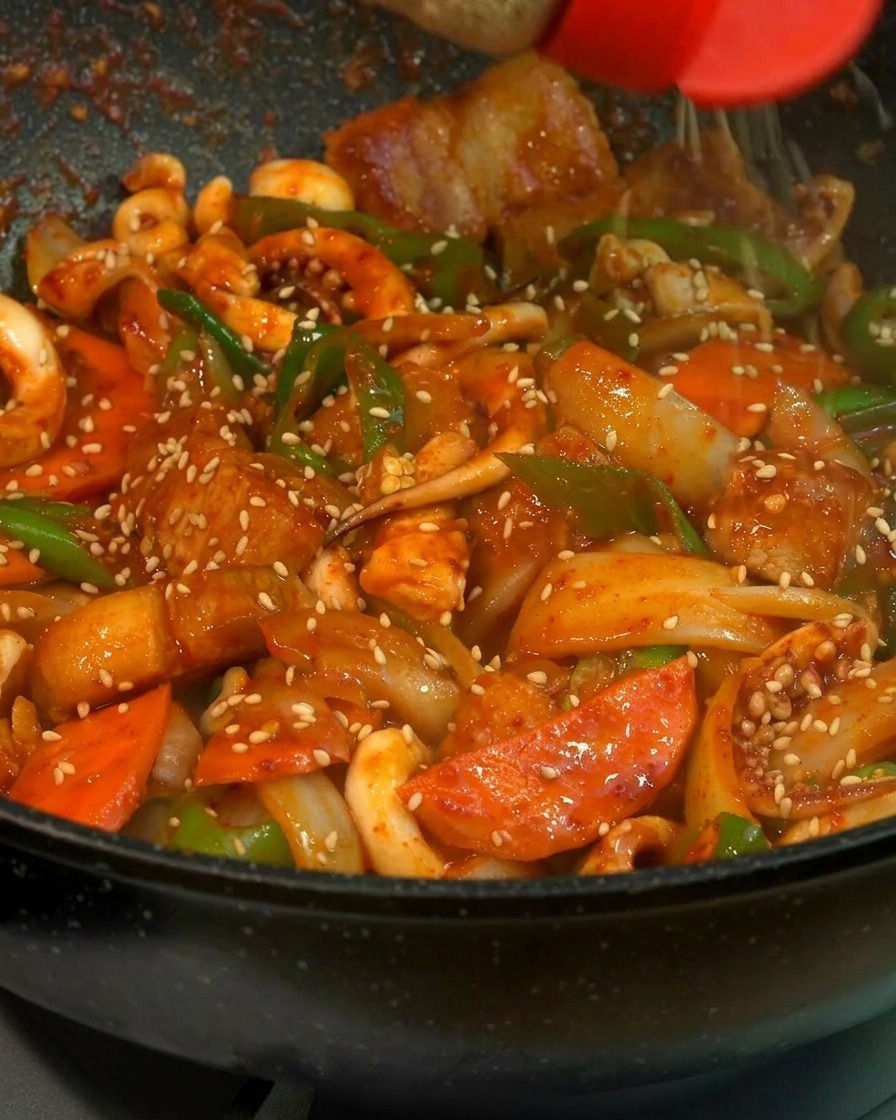 A close-up shot of white sesame seeds being sprinkled from a red-capped shaker onto a vibrant, glossy stir-fry of squid, pork belly, carrots, and green peppers in a black pan.