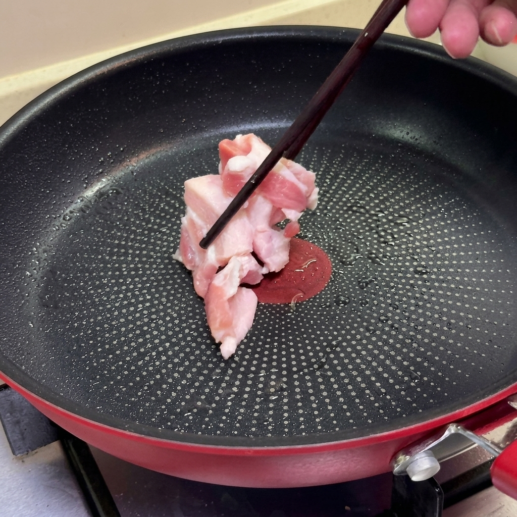 Chopsticks placing slices of raw pork belly into a heated non-stick frying pan.