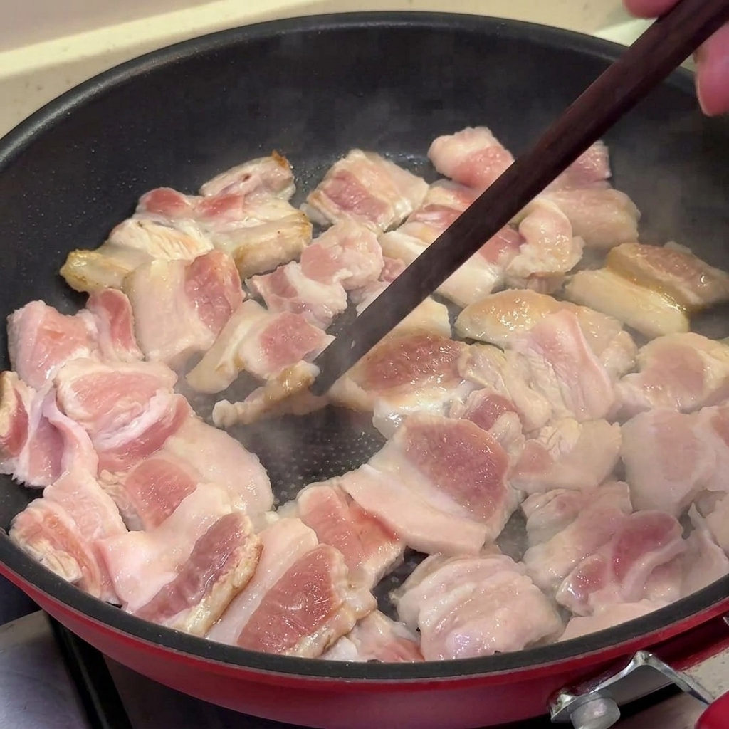 Slices of pork belly being pan-fried in a non-stick pan, turning golden brown and slightly charred, being stirred with chopsticks.