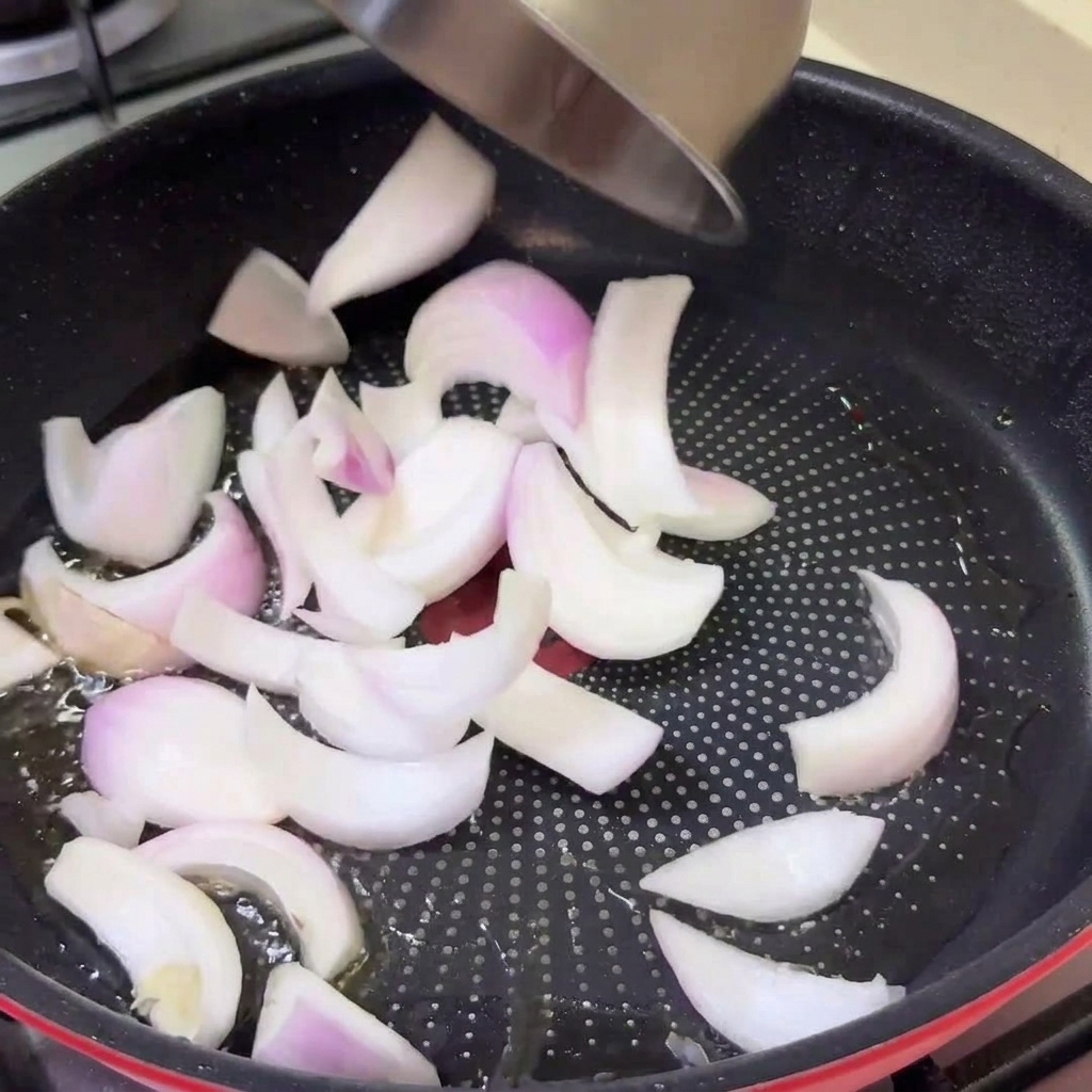 Sliced raw onions being poured from a bowl into a hot frying pan with oil.