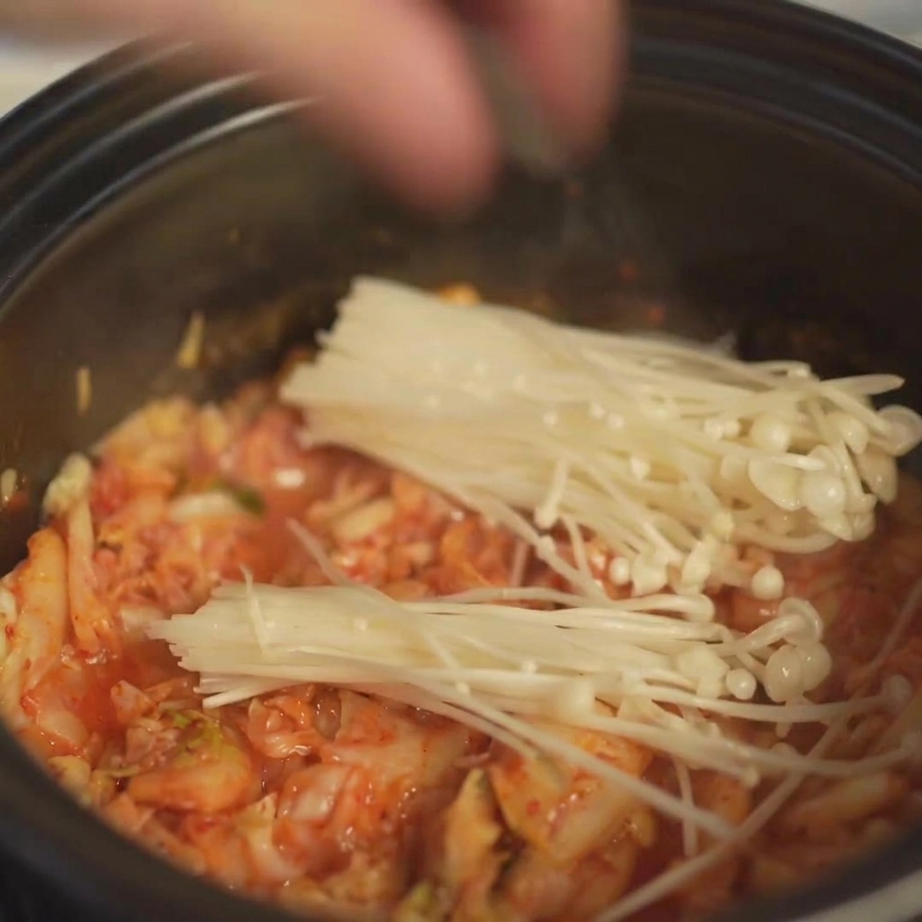 Placing a bundle of fresh enoki mushrooms into the pot over the kimchi.