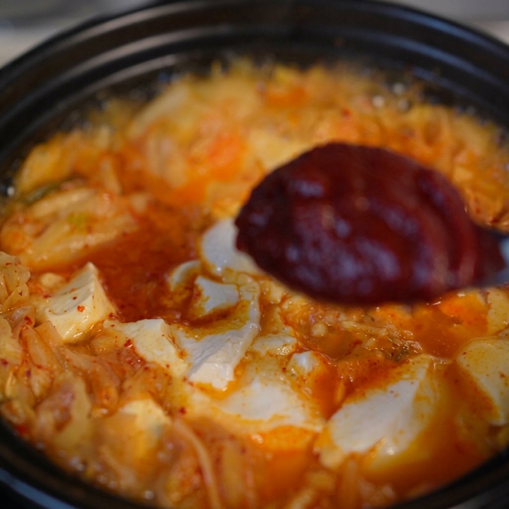 A spoon holding a dollop of thick, dark red Korean chili paste over a bubbling kimchi and tofu soup.