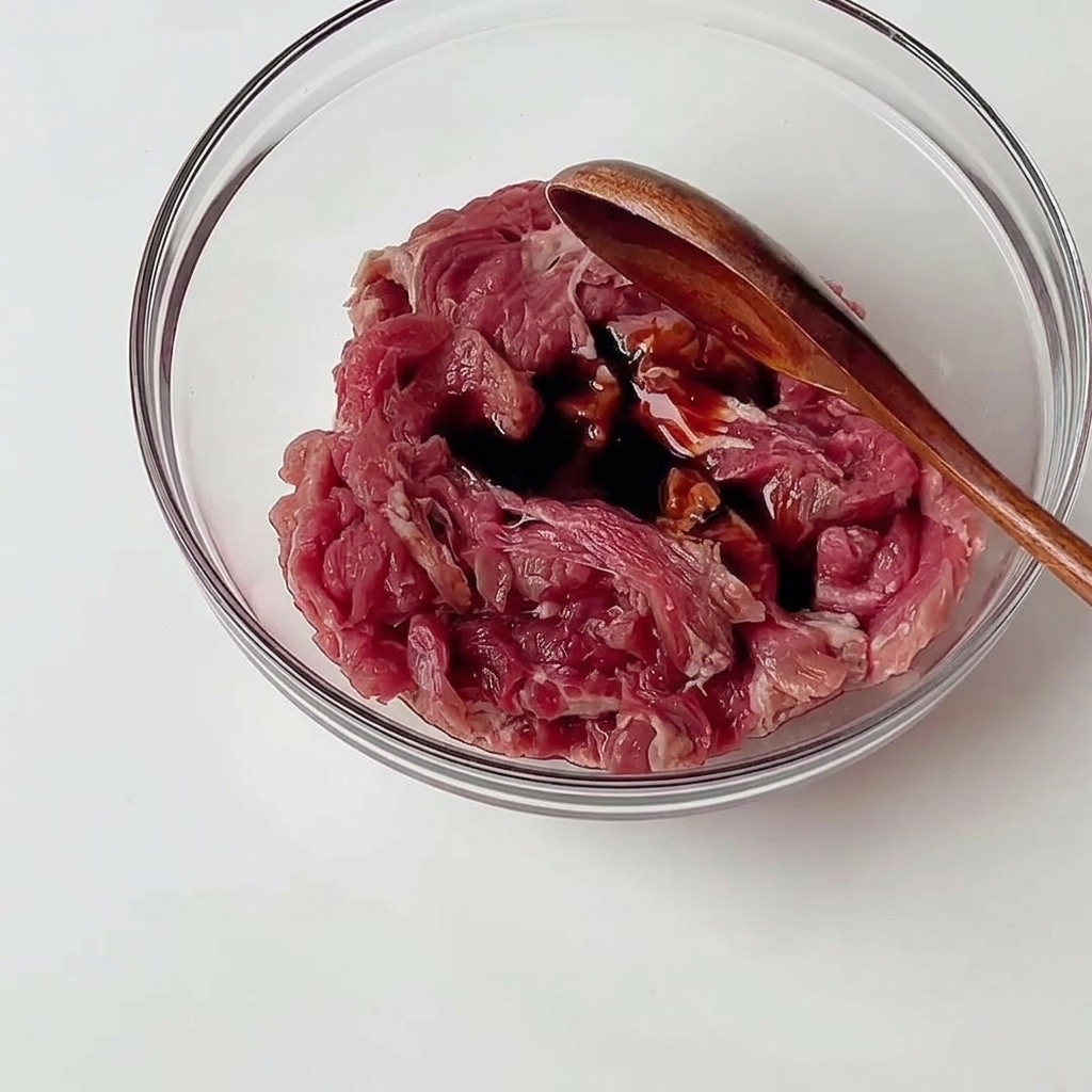 Thinly sliced raw beef in a clear glass bowl being mixed with dark savory sauces using a wooden spoon.