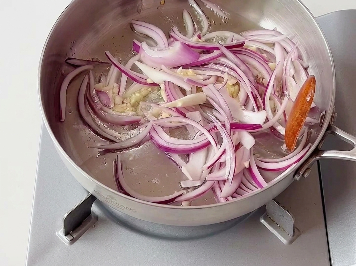 Sliced red onions and pieces of minced garlic sizzling in a shallow stainless steel pan on a portable stove.