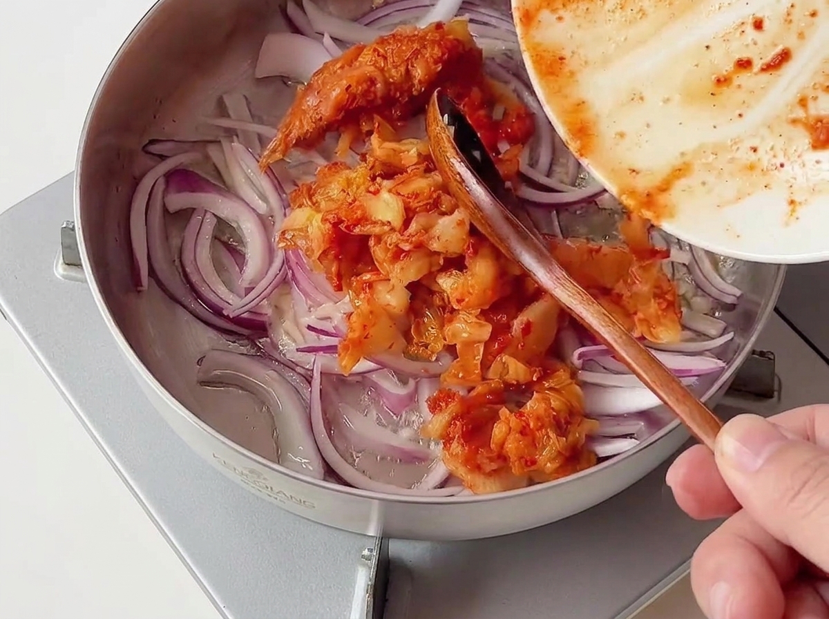 A portion of chopped red kimchi being added from a plate into a stainless steel pan containing sliced red onions.