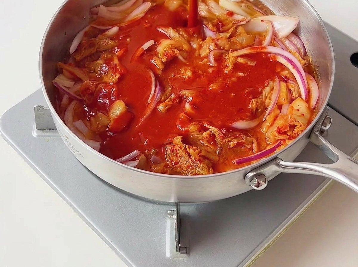 Bright red chili sauce being poured over sautéed kimchi and red onions cooking inside a silver pan.