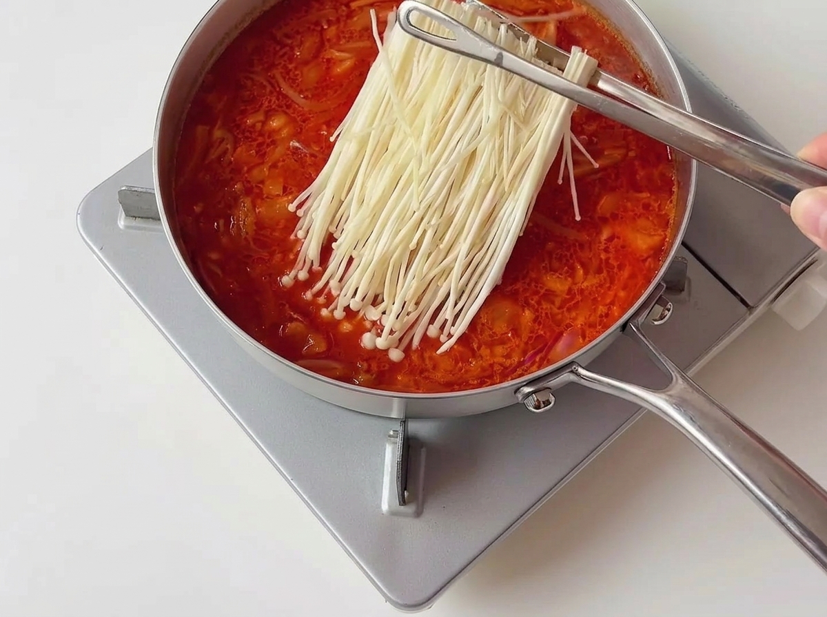 Metal tongs placing a neat cluster of white enoki mushrooms into a bubbling red kimchi stew.