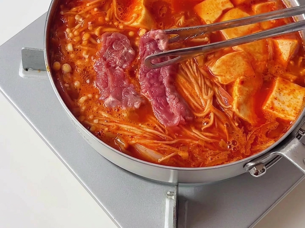 Tongs transferring pieces of pink, marinated raw beef into a hot, bubbling red stew next to tofu slices.
