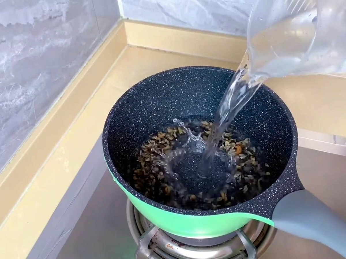 Clear water being poured from a plastic measuring cup into a green pot on a gas stove.