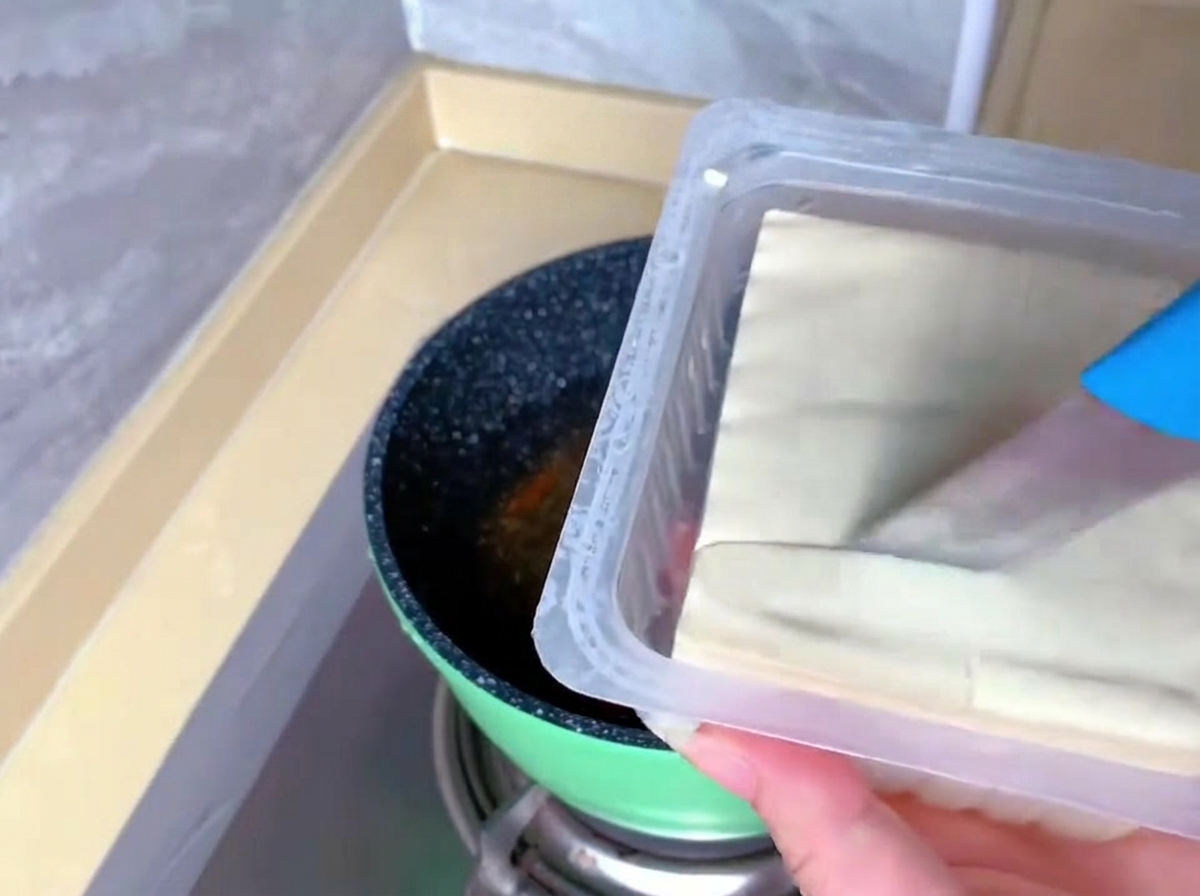 A person uses a blue knife to slice a block of white silken tofu directly inside its plastic packaging tray.