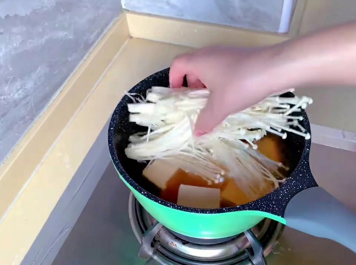 A hand transferring a bundle of long, white enoki mushrooms into a pot already containing broth and tofu.