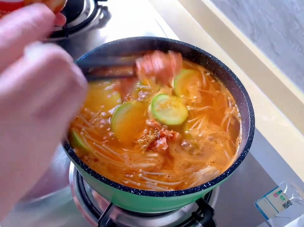 Adding pieces of red kimchi cabbage with chopsticks into a small green pot of bubbling orange soup with enoki mushrooms and round zucchini slices.