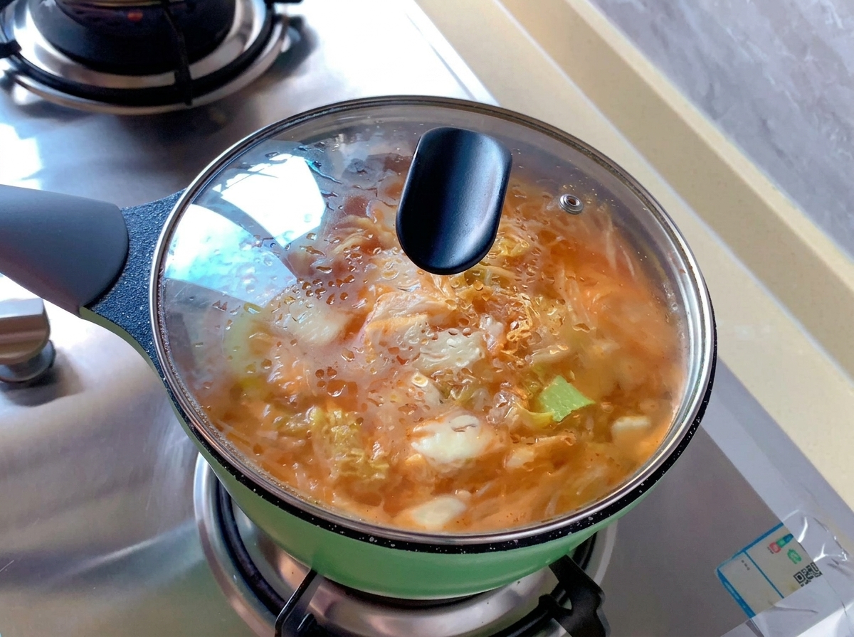 A small green pot filled with orange kimchi soup covered by a transparent glass lid with steam condensation on the surface.