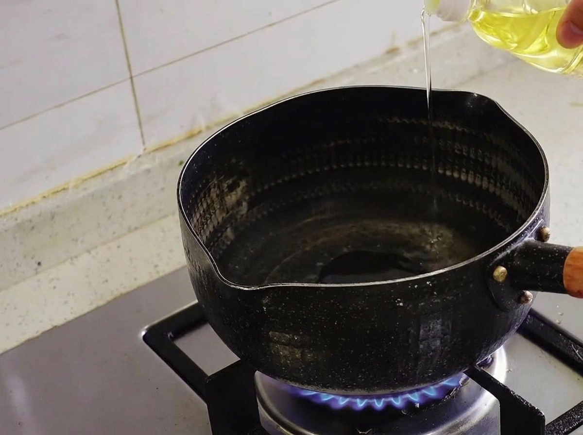 Clear oil being poured from a bottle into a black pot over a lit gas stove burner.