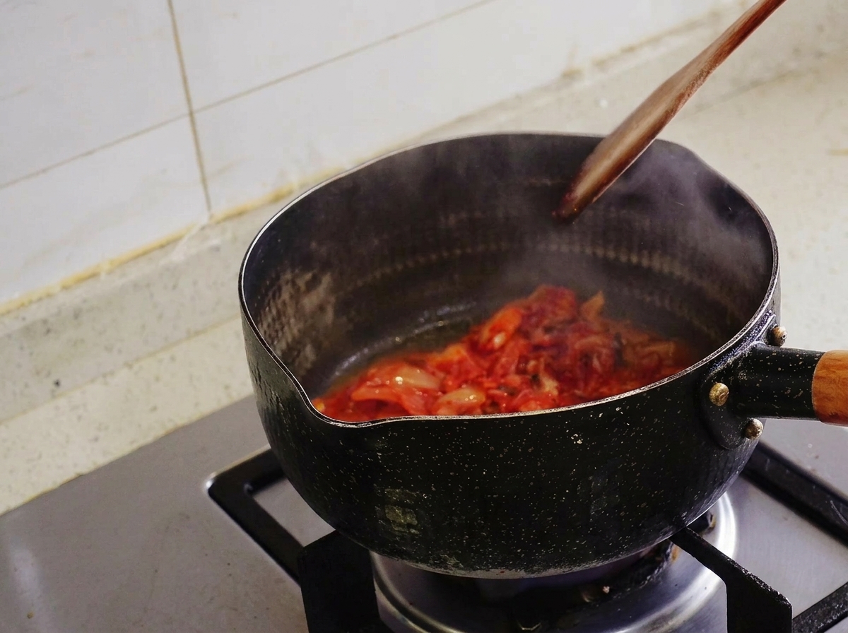 Kimchi being stir-fried with a wooden spatula inside a black pot, with steam rising.