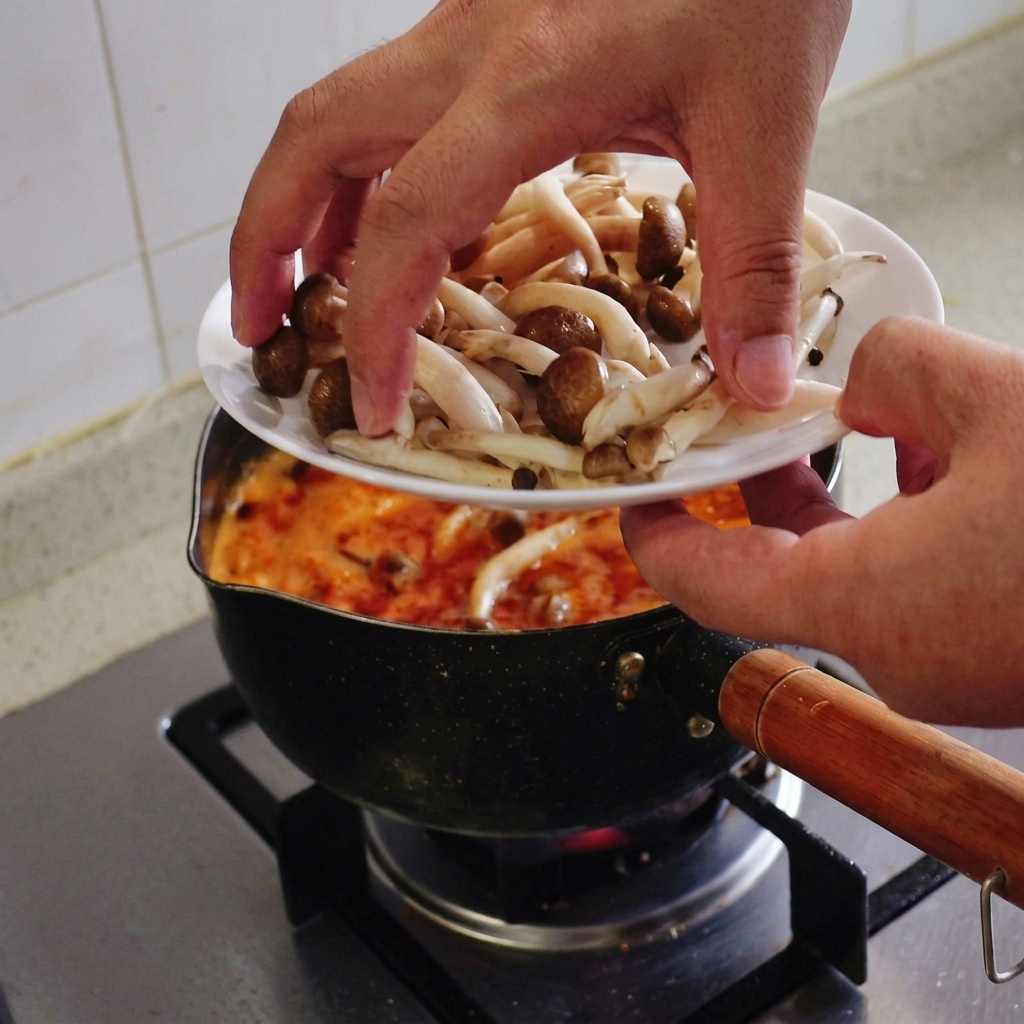 Hands transferring a handful of fresh brown-capped shimeji mushrooms from a white plate into a boiling pot of red soup.